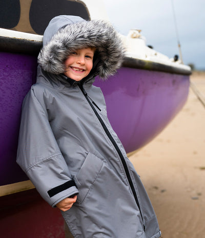 *MALE* A smiling child wearing a gray hooded Dryrobe with a faux-fur trimmed hood, standing beside a purple boat on a sandy beach.