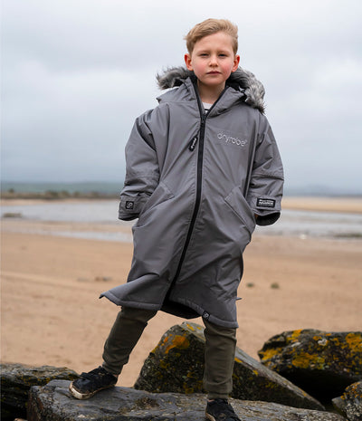 *MALE* A young boy wearing a gray dryrobe kids waterproof parka with a faux-fur trimmed hood, standing on rocks at a sandy beach under an overcast sky.