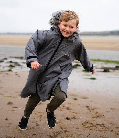 *MALE* A young boy smiling and running on a sandy beach wearing a gray waterproof Dryrobe with a faux-fur trimmed hood and olive green pants.