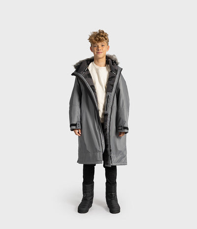 *MALE* A child model wearing a long gray hooded Dryrobe with dark faux-fur trim, a cream shirt, black pants, and black boots, standing against a light gray studio background.