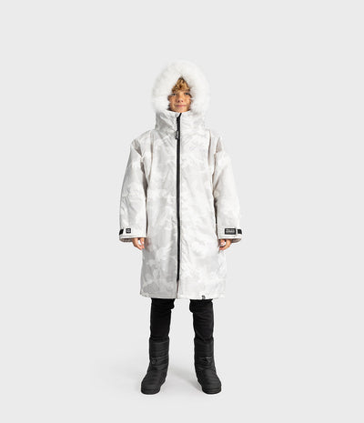 *MALE* A child wearing a long white hooded winter Dryrobe with a subtle camo pattern, white faux-fur trim around the hood, black pants, and black boots, standing against a light gray studio background.