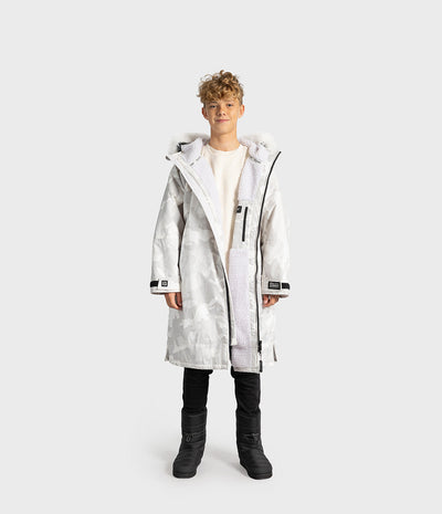 *MALE* A child model wearing a long white hooded Dryrobe with a subtle gray marbled pattern, black zippers, and a white sherpa lining, paired with a cream shirt, black pants, and black boots, set against a solid light gray background.