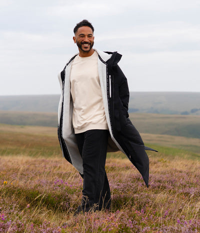 *MALE* A man standing in an open grassy field with purple flowers under an overcast sky, wearing a long black coat with a light gray interior lining over a cream sweatshirt and black trousers.
