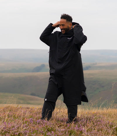 *MALE* A man wearing a black hooded rain jacket and matching black pants stands in a grassy field with rolling hills in the background under an overcast sky.