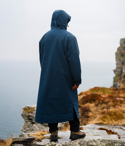 *MALE* A person viewed from behind wearing a long navy hooded raincoat while standing on a rocky cliffside overlooking water and grassy terrain.