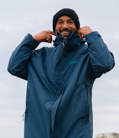 *MALE* A man wearing a blue Dryrobe waterproof jacket and a dark knit beanie, smiling and adjusting the collar of the jacket against an overcast sky. 