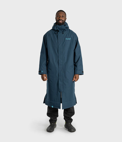 *MALE* A man wearing a long, dark teal-blue hooded raincoat with a small light blue 'Dryrobe' logo on the left chest, paired with black pants and dark boots, standing against a light grey studio background.