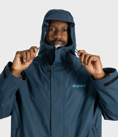*MALE* A man wearing a dark slate blue hooded Dryrobe jacket, holding the hood up with both hands against a light gray studio background.
