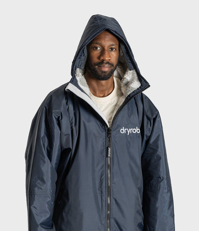 *MALE* A man wearing a navy blue hooded Dryrobe with a white sherpa lining and a white 'dryrobe' logo on the chest, photographed against a light gray studio background.
