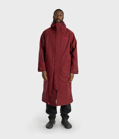 *MALE* A man wearing a long maroon hooded technical parka with a small pink logo on the left chest, paired with black pants and dark shoes, standing against a light gray studio background.