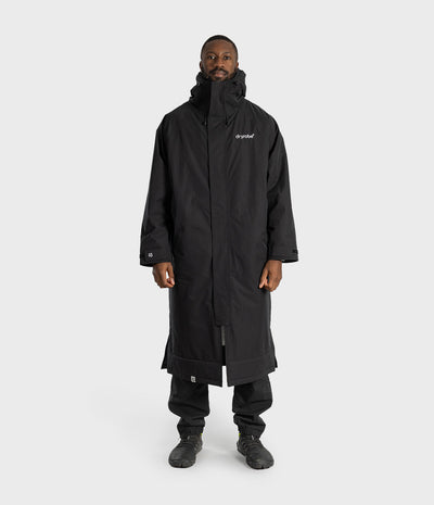 *MALE* A man wearing a long black hooded coat with a white 'branded' logo on the left chest, black pants, and black shoes, standing against a light gray studio background.