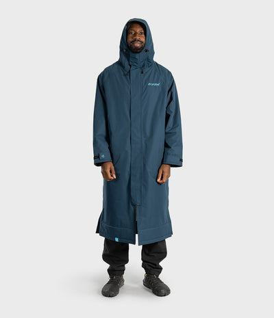 *MALE* A man wearing a long, hooded waterproof coat in a muted blue-gray color, paired with black pants and boots, standing against a light gray studio background. The brand name 'Dryrobe' is visible on the left chest of the coat.