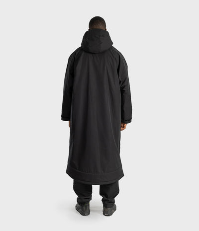 *MALE* A person viewed from the back wearing a long black hooded coat over black pants and dark shoes, standing against a plain light gray studio background.