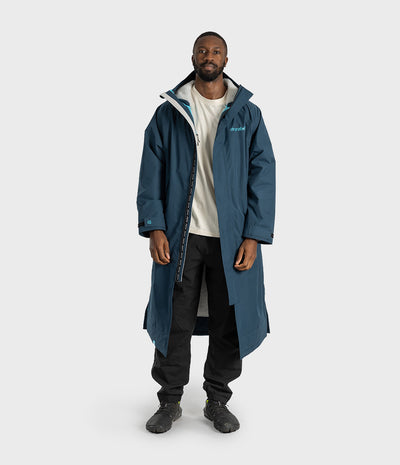 *MALE* A man modeling a long, dark blue hooded jacket over a cream t-shirt, black pants, and black shoes, standing against a light gray studio background.