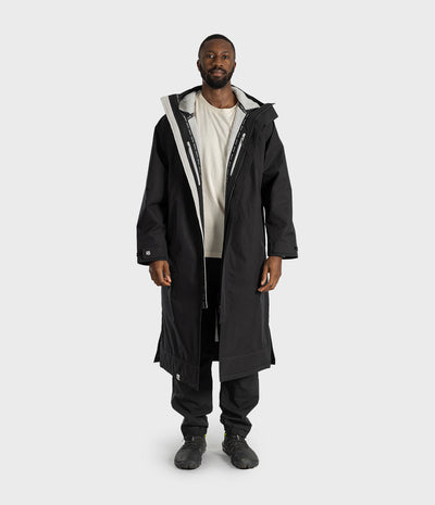 *MALE* A man modeling a long black hooded parka with a white inner lining and silver zipper details, worn over a white t-shirt, black pants, and black sneakers, against a light gray studio background.