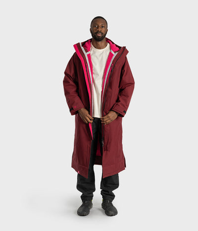 *MALE* A man wearing a long burgundy hooded parka with a bright pink inner lining and zipper trim, layered over a white t-shirt, black pants, and black boots, standing against a light gray studio background.
