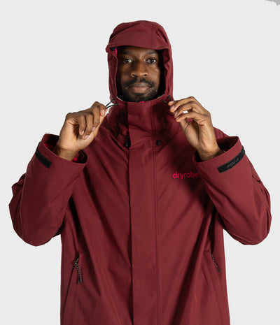 *MALE* A man wearing a maroon hooded Dryrobe jacket, holding the hood up. The jacket has a small pink 'dryrobe' logo on the left chest and black trim details on the cuffs.