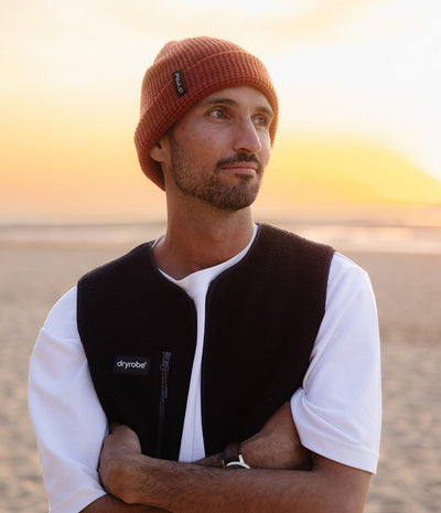 A man wearing a rust-colored knit beanie and a black Dryrobe fleece vest over a white t-shirt, standing on a sandy beach at sunset.