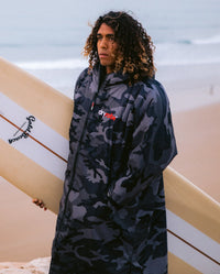 *MALE* standing on a beach holding a longboard, wearing Black Camo dryrobe® Advance Long Sleeve