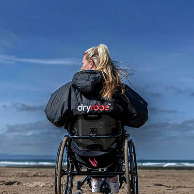 Person in a wheelchair wearing a black Adapt jacket with Dryrobe® branding on a beach.
