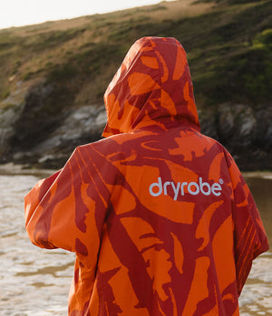 *MALE* A person standing outdoors by the water, shown from behind wearing a hooded Dryrobe rain jacket with an orange and red abstract camouflage pattern and the Dryrobe logo printed in white on the back.