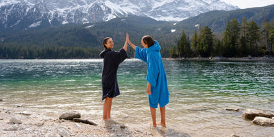 Two people in towel robes giving each other a high-five by a lake with mountains in the background