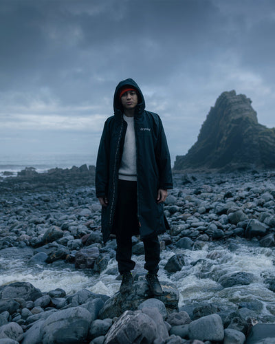 *MALE*Person wearing a dark coat standing on a rocky beach with a stormy sky.