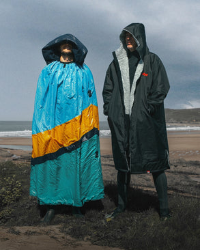 Two people wearing oversized hooded changing robes by the beach, one in a turquoise, yellow, and navy robe, the other in a black robe with a grey fleece lining, standing on grassy cliffs with the sea in the background.