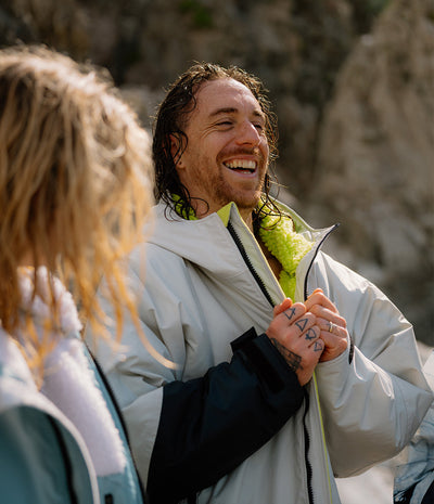 *MALE* Two people standing outdoors, one with tattoos on hands and arms, wearing light-colored Dryrobes. 