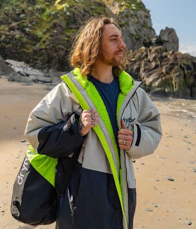 *MALE* Man standing on a beach wearing a Dryrobe with a visible brand logo, holding a bag.