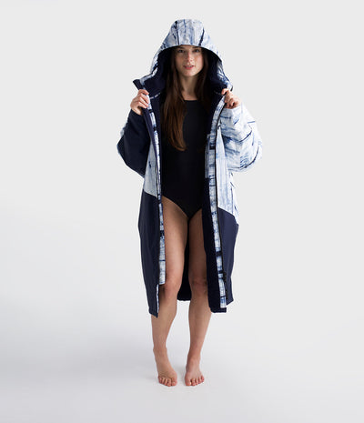 Woman wearing a black swimsuit and a long, dark blue hooded Dryrobe with white patterns on a white background