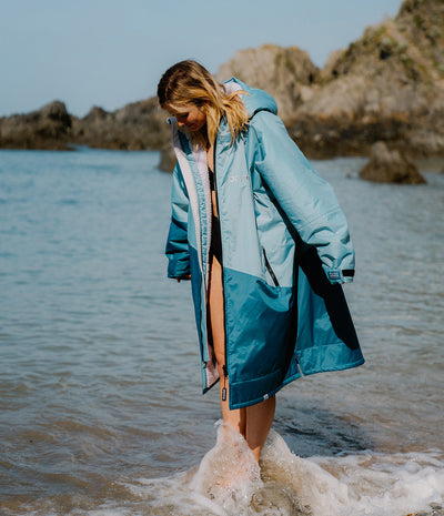 Person wearing a blue Dryrobe standing in shallow water near rocks.