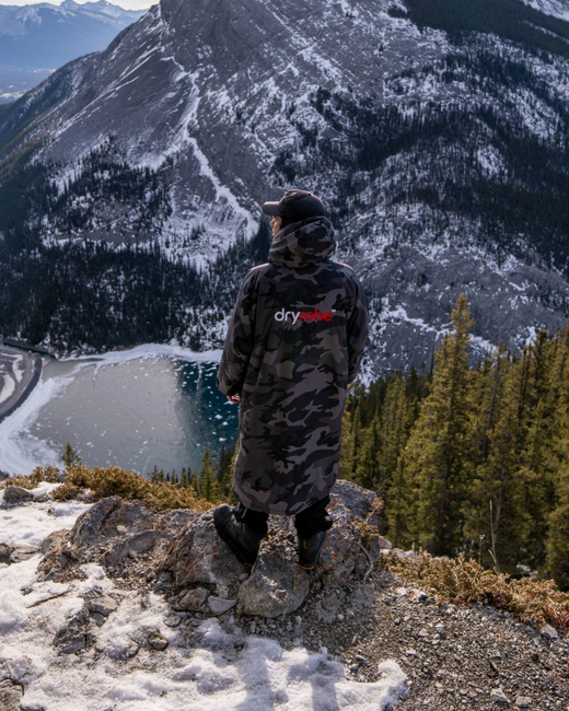 Person wearing a camouflage Dryrobe standing on a mountain overlooking a lake and snow-covered mountains.