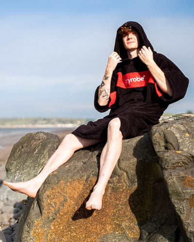 Man sat on rocks on a beach wearing Organic Towel dryrobe Remix in Black Red