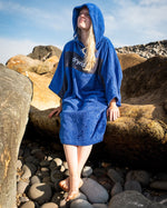 Woman sat on rocks on a beach wearing Organic Towel dryrobe Remix in Royal Blue Navy with hood up