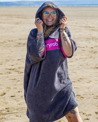Woman smiling wearing sunglasses on a beach wearing Organic Towel dryrobe Remix in Slate Grey Pink with hood up 
