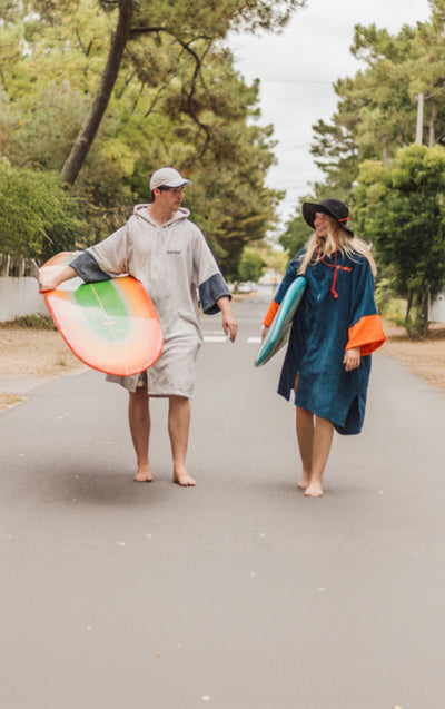 Two people walking on a road holding surfboards, surrounded by trees.