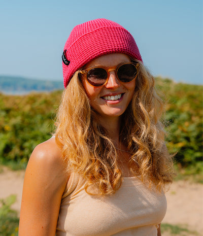 A smiling woman outdoors wearing a bright pink knit beanie, round sunglasses, and a beige ribbed tank top, with blurred greenery and blue sky in the background.