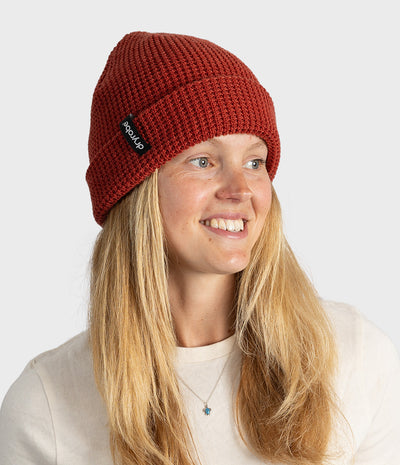 A smiling blonde woman wearing a rust-red ribbed knit beanie with a small black brand tag that reads 'dryrobe'. The photo is taken against a clean, light gray studio background.