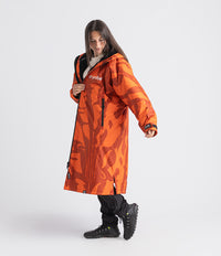 A woman modeling a bright orange hooded parka with a dark abstract camouflage-like pattern, black zippers, and the brand name 'dryrobe' printed on the chest, set against a plain light gray studio background.