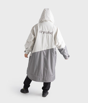 A person shown from the back wearing a hooded Dryrobe rain jacket in off-white and gray, with black pants and shoes, against a light gray studio background.