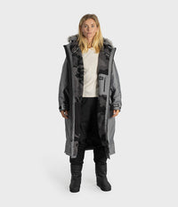 A woman modeling a long gray hooded dryrobe with faux-fur trim, worn over a cream top, black patterned jacket, black pants, and black boots, against a light gray studio background.