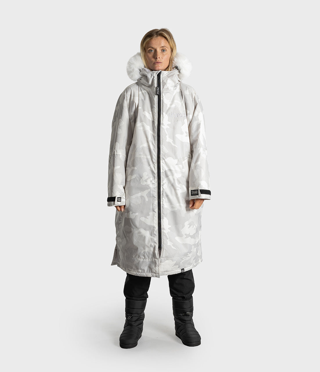 A woman modeling a long hooded winter Dryrobe with a light gray and white camouflage pattern, black zipper, and a fluffy white fur-trimmed hood, paired with black pants and boots against a light gray studio background.