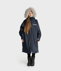 A young girl wearing a dark navy mid-length waterproof Dryrobe with a white 'dryrobe' logo on the chest, a gray faux-fur trimmed hood, black pants, and black boots, standing against a light gray studio background.