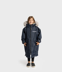 A young girl stands against a light gray studio background wearing a navy blue hooded Dryrobe with a faux-fur trim and the brand name 'dryobe' printed on the chest.