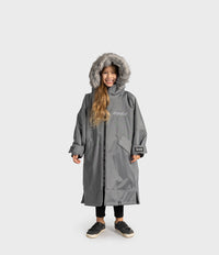 A child wearing a long gray hooded Dryrobe with a faux-fur trimmed hood, front zipper, and flap pockets, standing against a light gray studio background. The brand name 'dryrobe' is printed on the chest.
