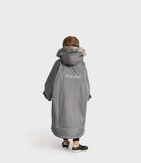 A child viewed from behind wearing a long gray hooded Dryrobe with a faux-fur trim and the brand name 'dryrobe' printed on the back, against a light gray studio background.