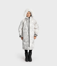 A child wearing a long, white hooded winter Dryrobe with a white faux-fur trim around the hood, black pants, and black boots, standing against a light gray studio background.