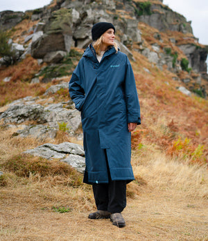 A woman wearing a long navy blue waterproof coat, black beanie, and black trousers stands outdoors in a rocky, grassy mountain setting. The brand name 'outward' is visible on the left chest of the coat.