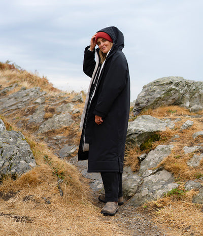 A person standing outdoors on a rocky hillside wearing a long black hooded coat, dark pants, brown boots, a red beanie, and a light scarf.